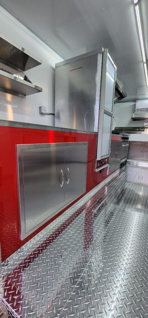 Interior of a custom BBQ food trailer showing a stainless steel dual electric food warmer, aluminum diamond plate flooring, and storage cabinetry with stainless steel doors.